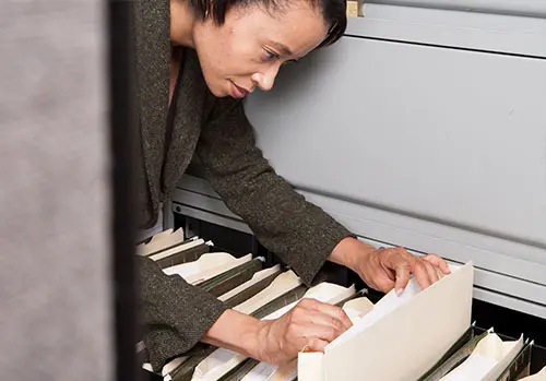 A business person stoops to examine the contents of a filing cabinet drawer.