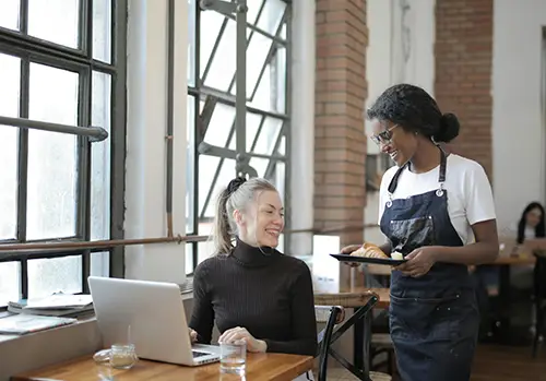 A woman sits at a table with her laptop and is served by a waitress.